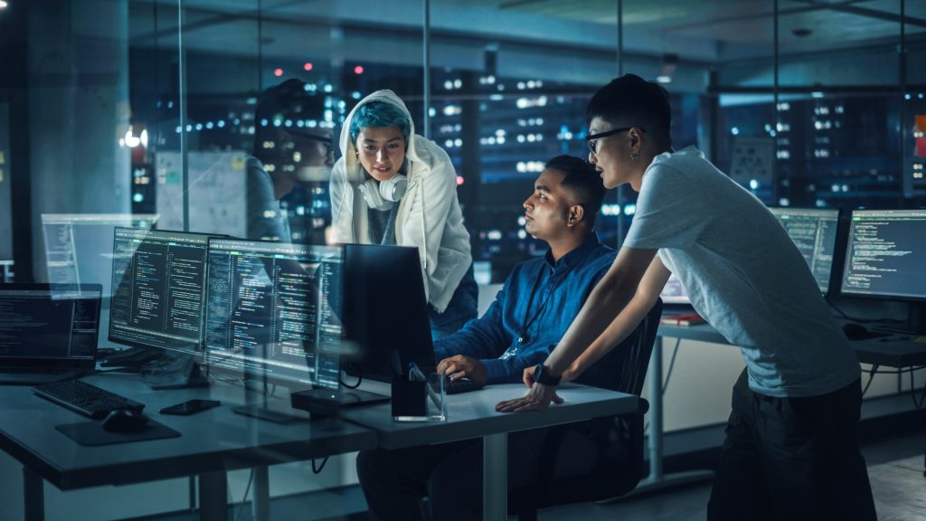 A security engineer in a dark office reviewing code on a laptop with a server rack visible in the background