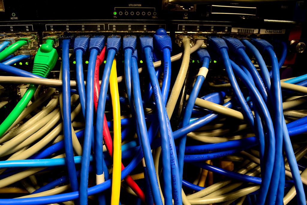 A developer in a dim server room watches a wall of blinking network lights while holding a coffee mug, stacks of backup drives on a metal shelf behind them.